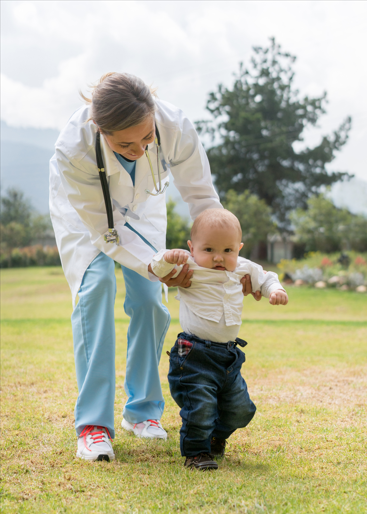 A woman in scrubs, white coat, and stethoscope walks outside, holding her baby in front of her. 
